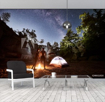 Picture of Back view silhouettes of young couple hikers man and woman having a rest beside camp bonfire and illuminated tourist tent in mountains enjoying beautiful night starry sky full of stars and Milky way